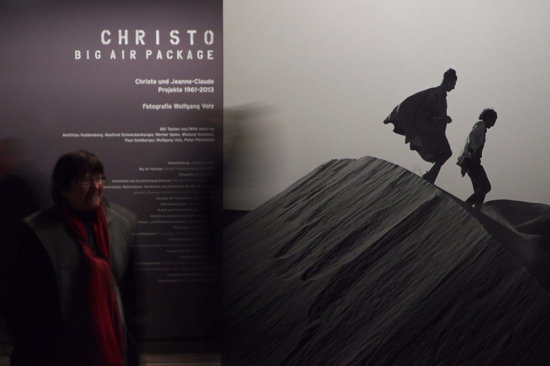 Eine Person vor einer Tafel mit einem grauen Bild von zwei Personen auf einer Düne und linksstehendem Text über CHRISTO – BIG AIR PACKAGE