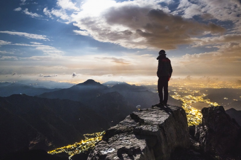 Foto von einem Wanderer auf einem Felsen auf einer Bergspitze mit Blick auf andere Berge und Lichter einer Stadt bei Dämmerung.